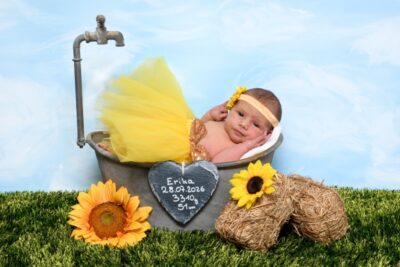 Die kleine Erika hat ein gelbes Tu Tu an und liegt in einer grauen Zinkwanne, davor liegen gelbe Sonnenblumen.