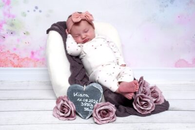 Die kleine Kiara liegt in einem weißen Sessel, sie hat ein rosa Stirnband auf und vor ihr liegen Rosen.
